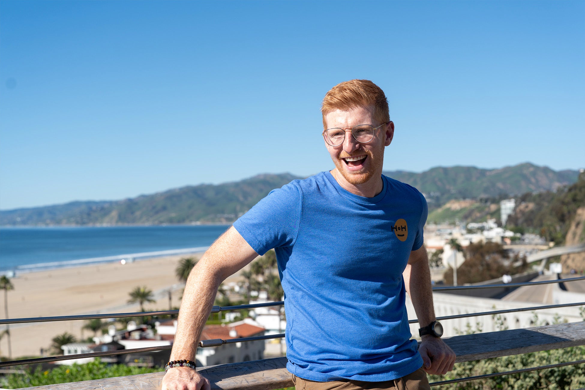 A smiling man wearing glasses and a blue T-shirt leans against a railing overlooking Los Angeles’ sunny beach and coastal hills.