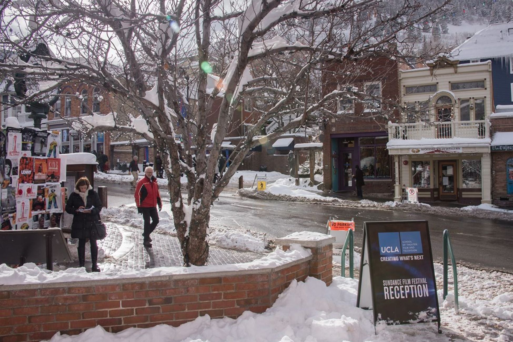 Snowy street scene with a "UCLA School of Theater, Film and Television" sign advertising a Sundance Film Festival reception, pedestrians walking past shops in the background.