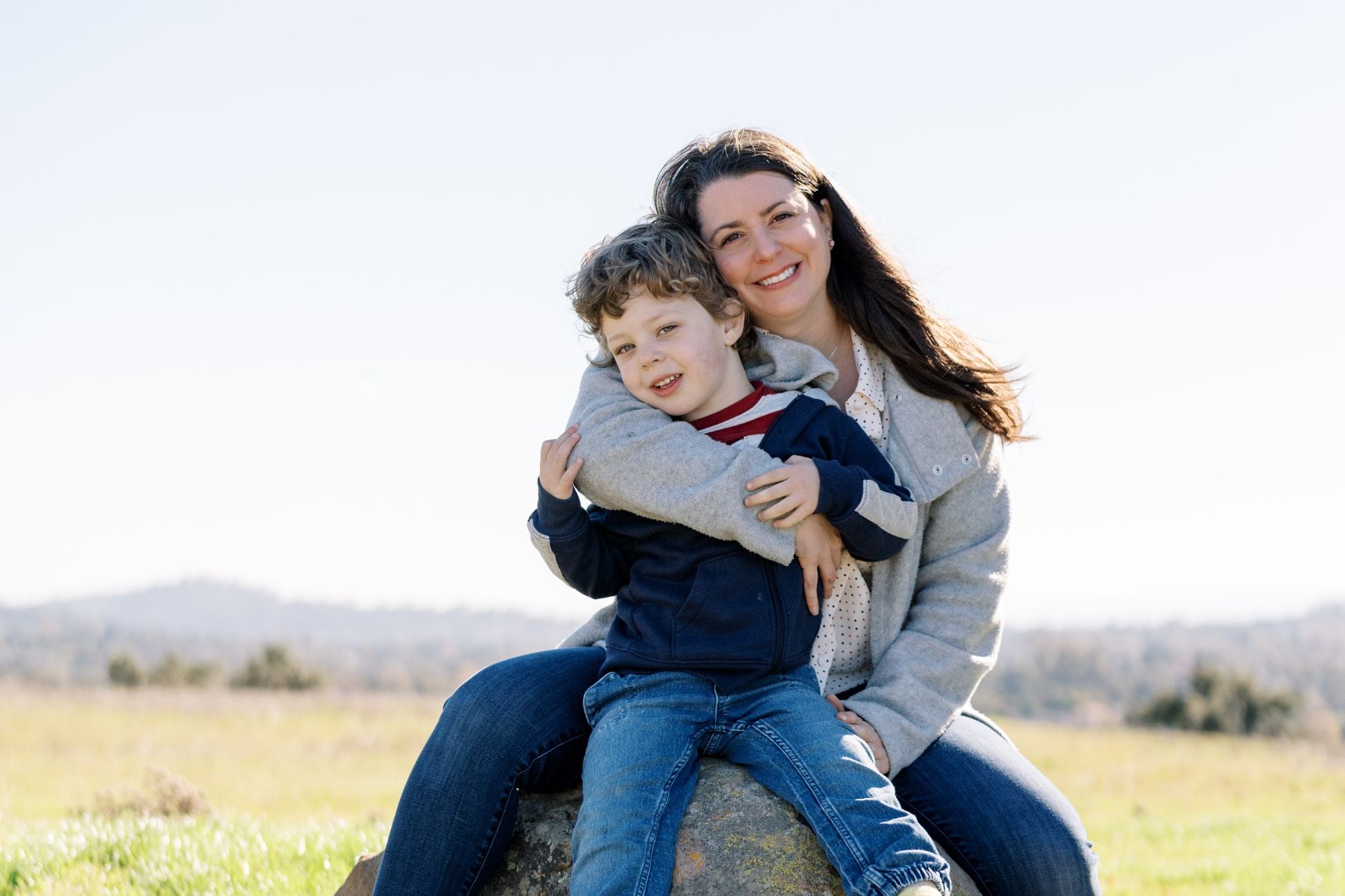 A woman hugs her son in a grassy field. Woman wears a gray sweater, jeans and long brown hair. Son wears a blue sweater, jeans, and light brown hair.