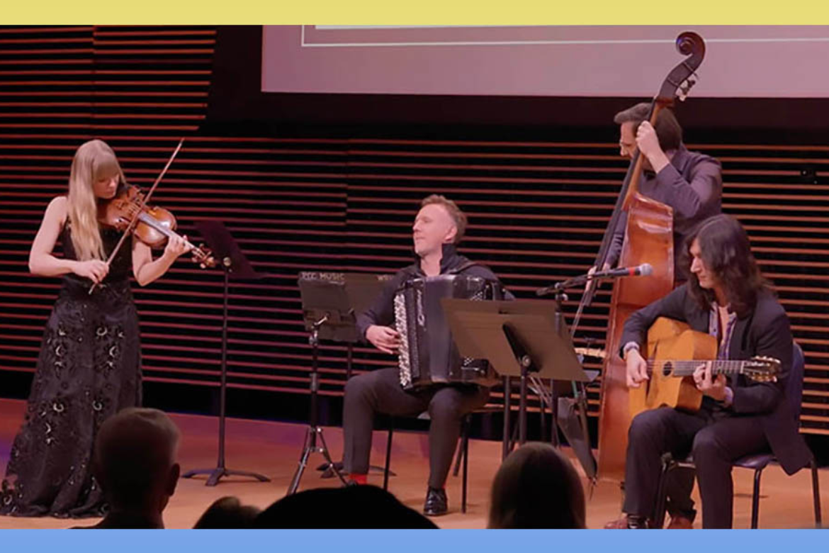 A four-piece ensemble performs onstage, featuring violin, accordion, double bass, and guitar, with music stands in front of the musicians and an audience visible in the foreground.