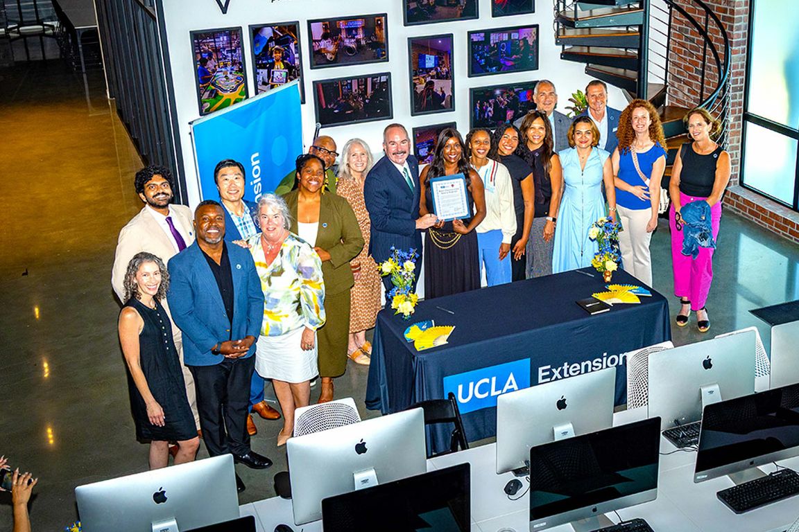 A group of people posing behind a table with a UCLA Extension banner, one holding a framed certificate, in a room with a photo wall and computer workstations in the foreground.