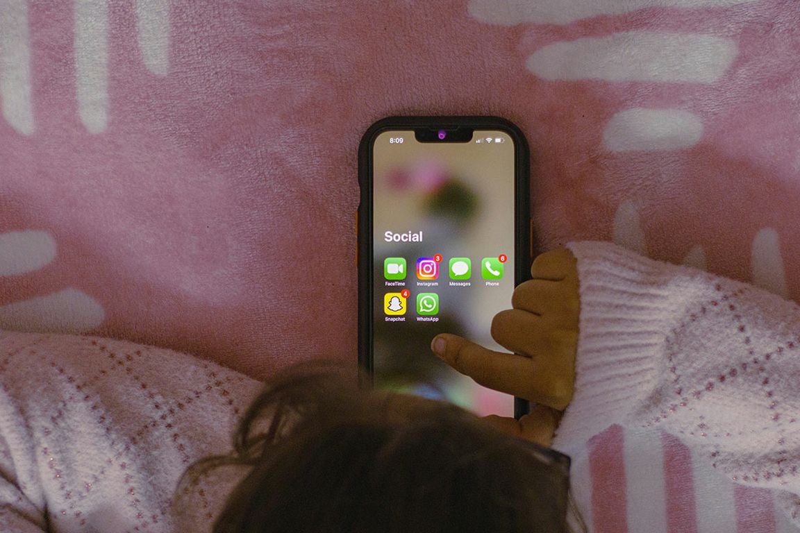 Child holding a smartphone displaying a social media folder with messaging and social apps against a pink background, suggesting mobile communication and social media use.